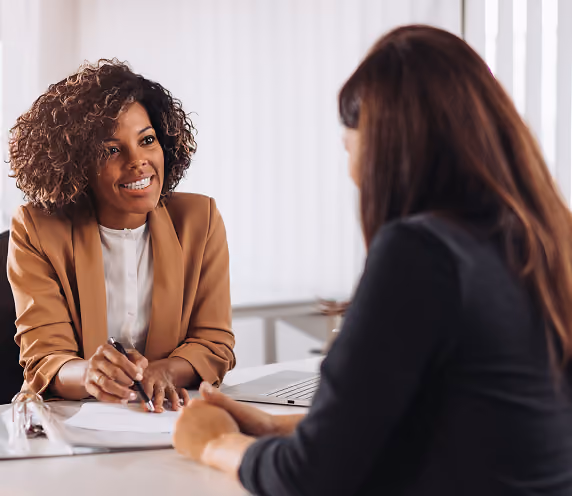 Two women in a professional setting engaged in a consultation or meeting, one with curly hair and a brown blazer smiling while holding a pen and paper, the other with straight dark hair listening attentively.