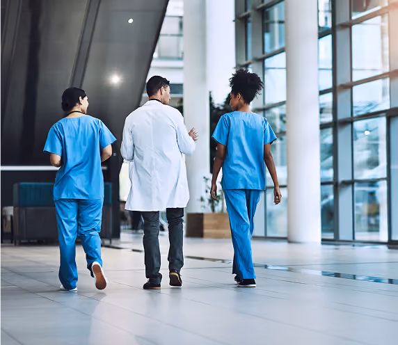 Two medical professionals in blue scrubs walk alongside a doctor in a white coat through a modern hospital corridor with large windows.