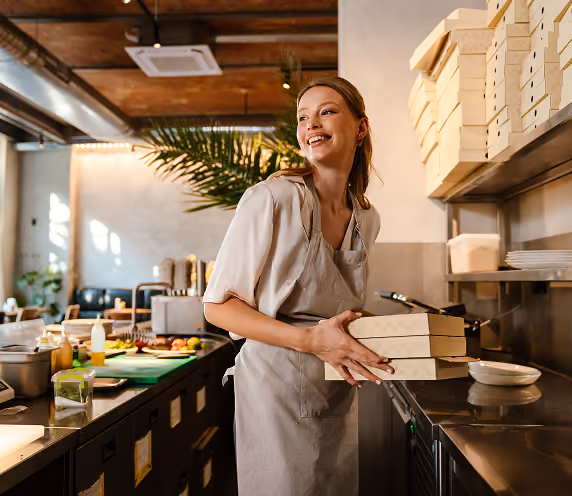 Smiling female food service worker in a beige apron holding stacked pizza boxes in a modern kitchen with wooden ceiling and shelves.