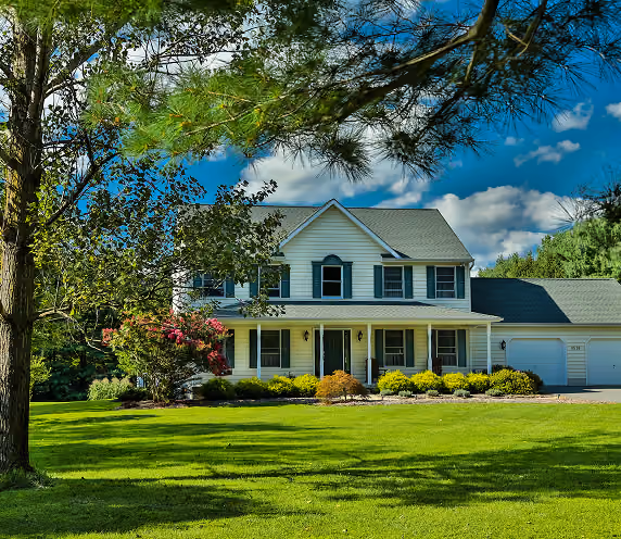 Two-story white single-family house with gray roof, front porch, and attached garage, surrounded by green lawn and trees under a partly cloudy blue sky.