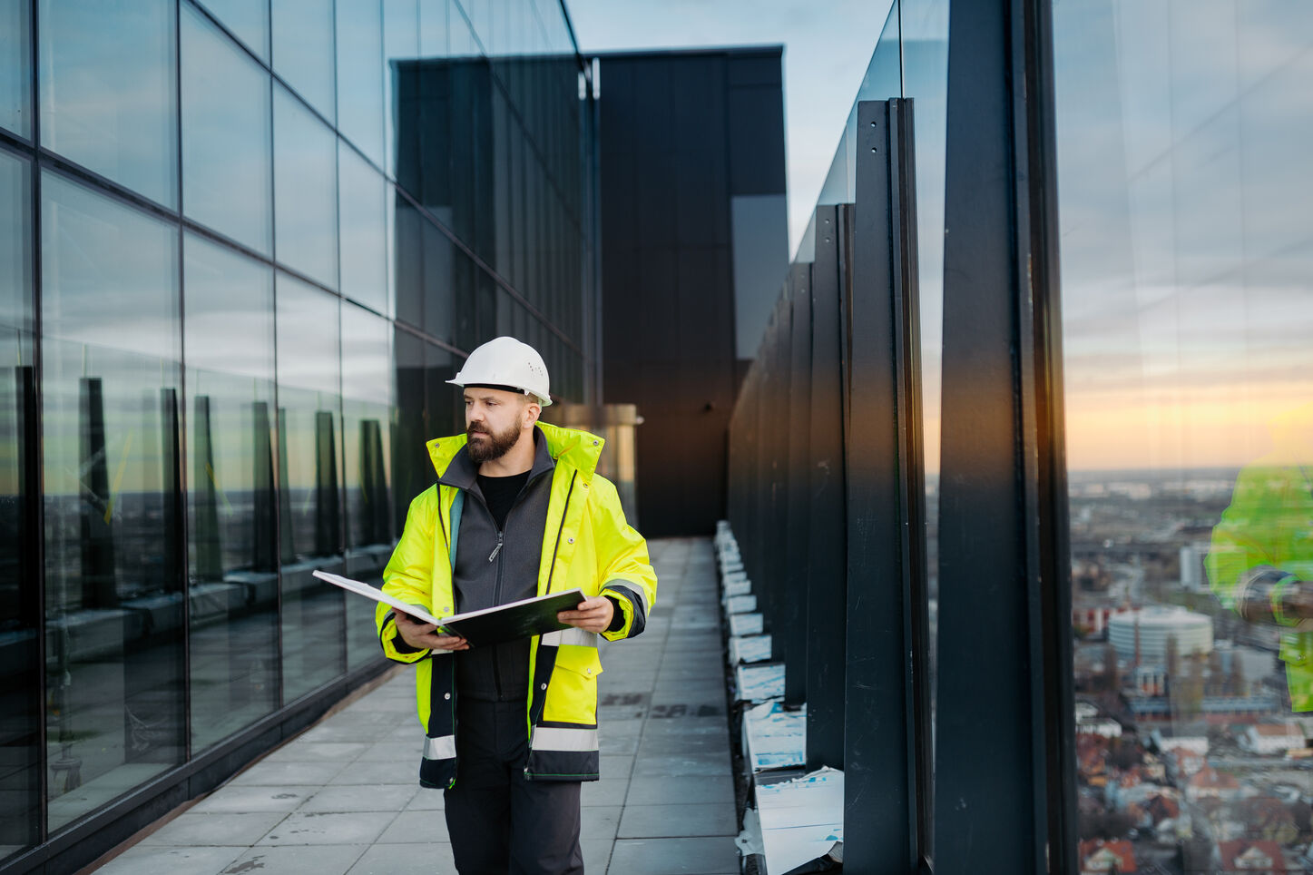 Man in a white construction helmet and yellow safety jacket holding an open binder, walking along a rooftop terrace next to a building with large glass windows reflecting the cityscape at sunset.