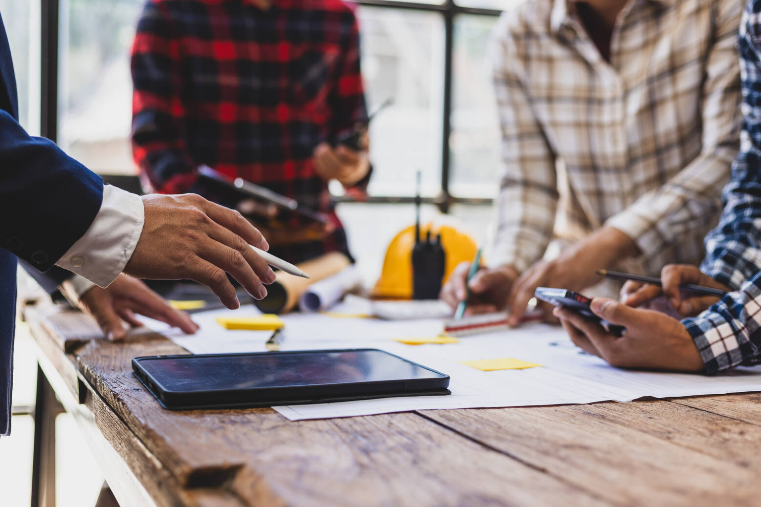 Close-up of a group of people collaborating over a wooden table with blueprints, a tablet, a yellow hard hat, and walkie-talkie, with hands holding pens and a calculator.
