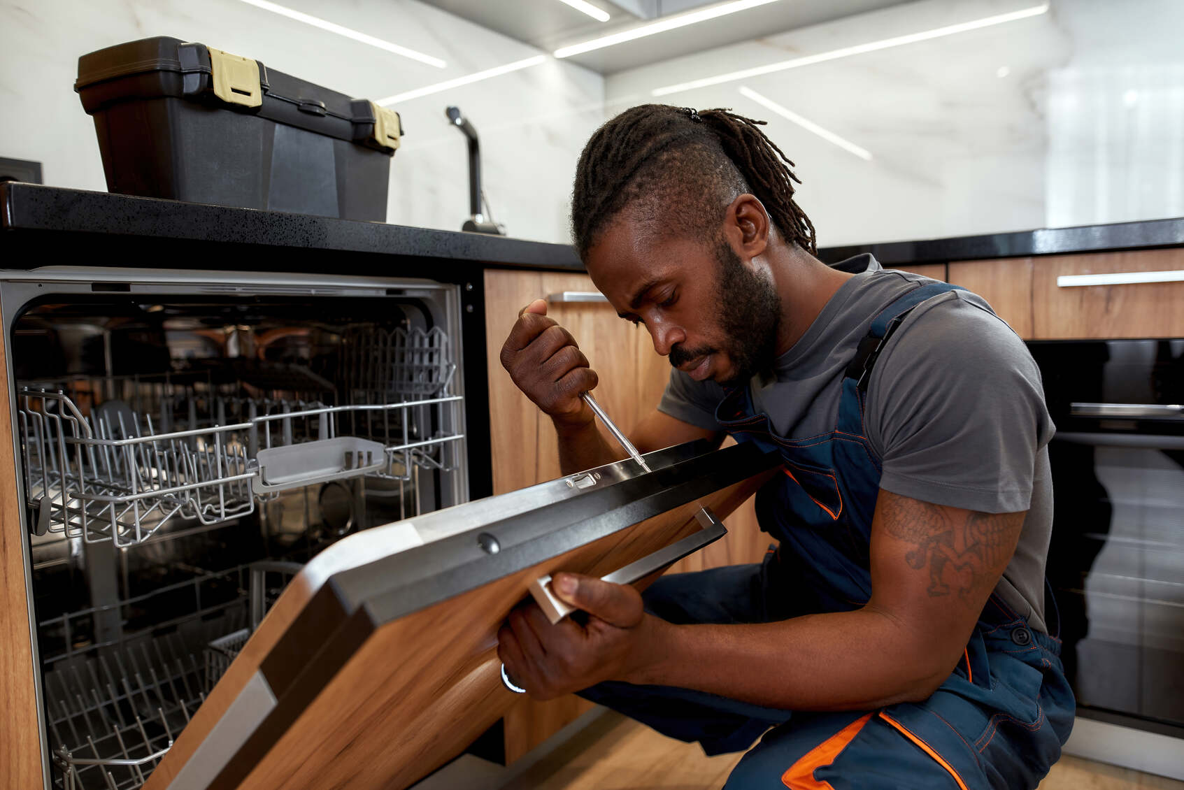 Man with dreadlocks and tattoo on arm wearing overalls uses a screwdriver to repair a dishwasher in a modern kitchen with black countertops and wooden cabinets.