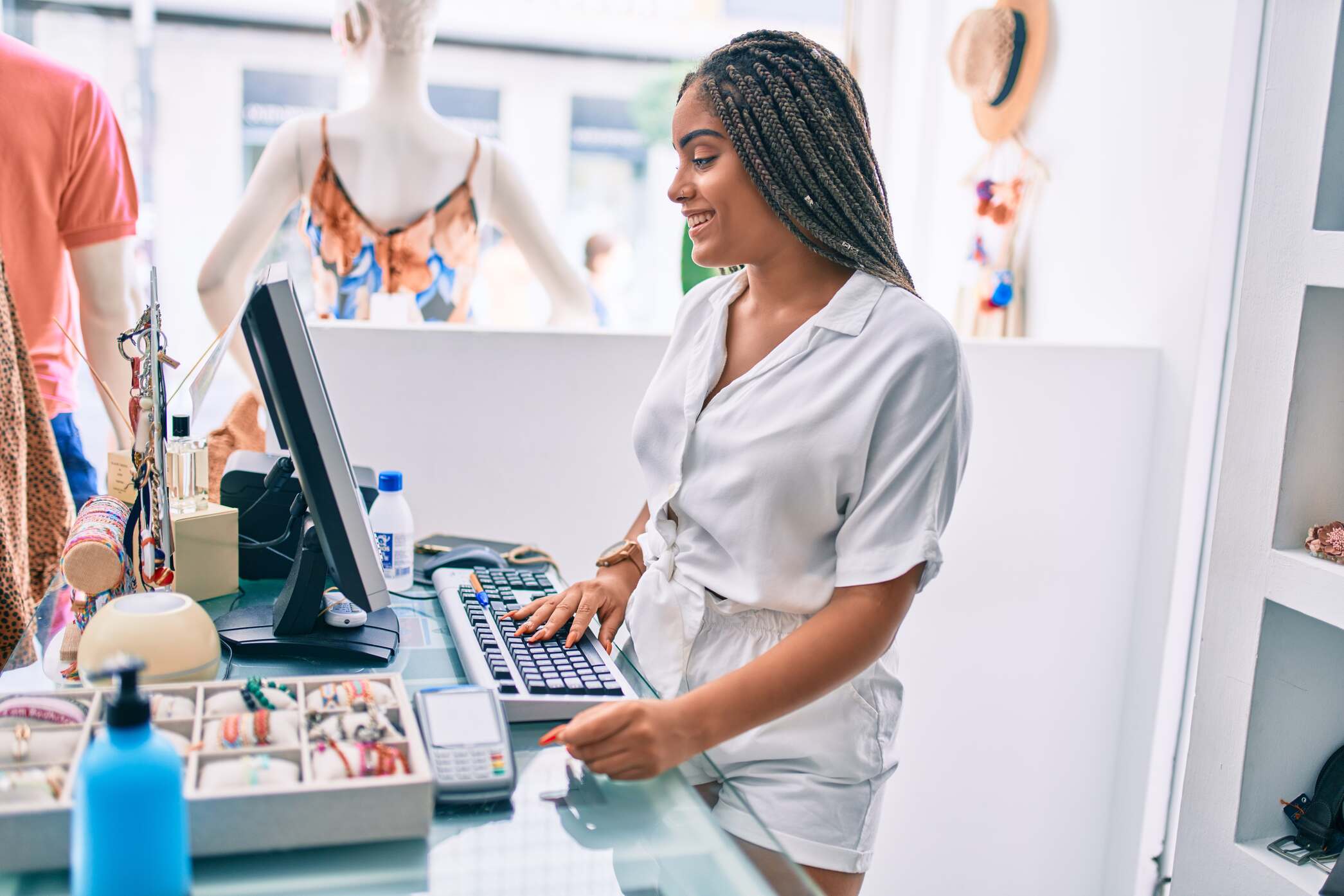 Smiling woman with braided hair wearing a white blouse and shorts, working at a retail cashier counter with computer, payment terminal, and jewelry display.