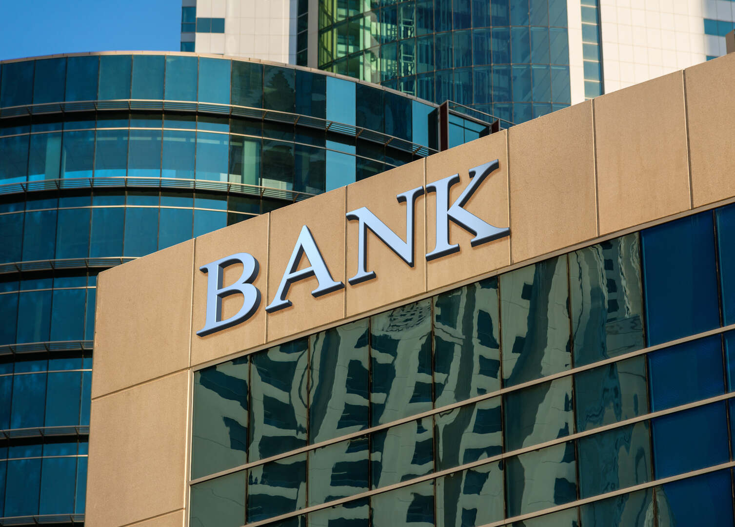 Close-up of a beige building facade with the word 'BANK' in large white letters above reflective blue-tinted windows showing distorted reflections of surrounding skyscrapers.