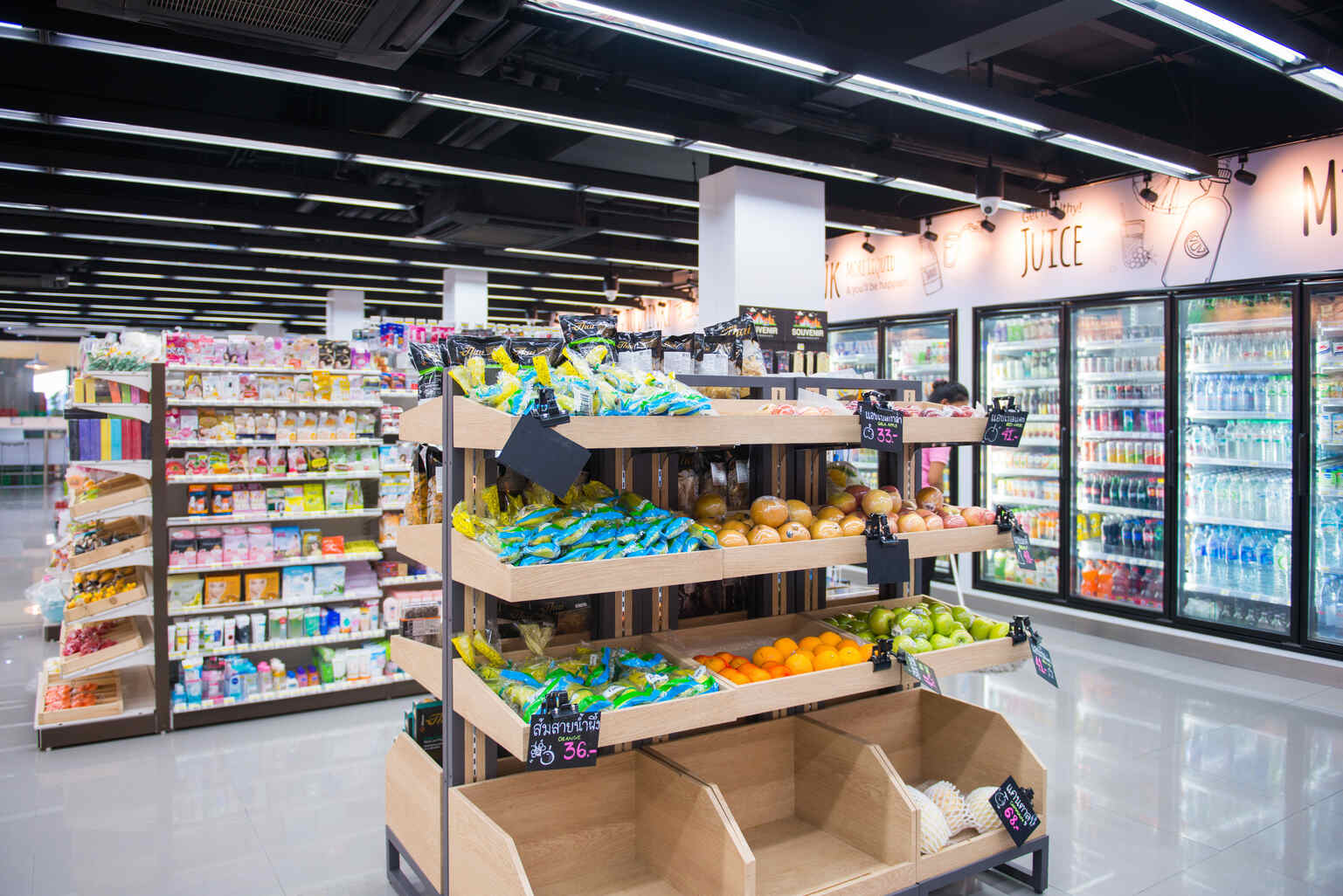 Supermarket interior showing wooden display shelves with various packaged snacks and fresh fruits including oranges and green apples, with refrigerated beverage coolers in the background.