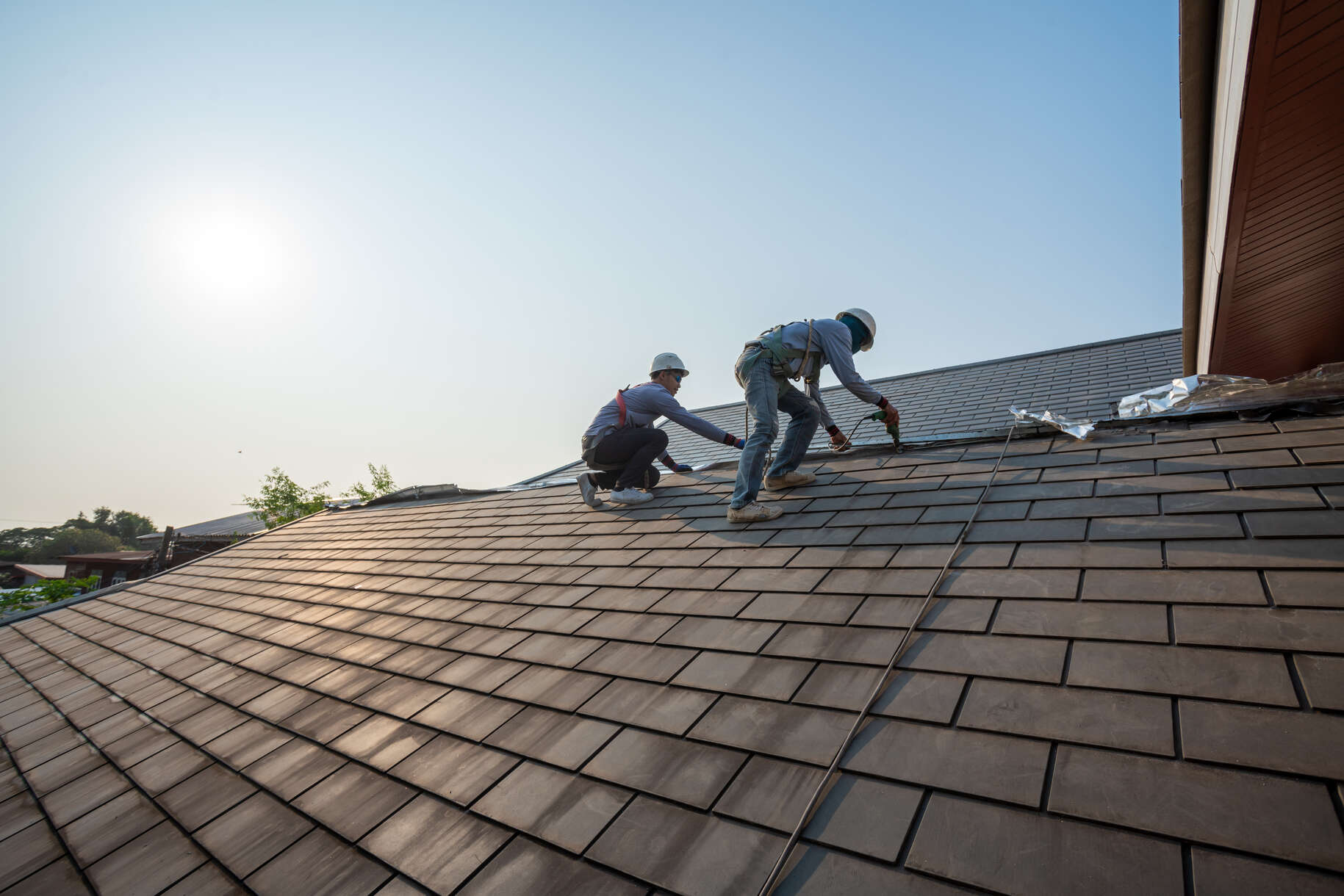 Two construction workers wearing safety harnesses and helmets installing or repairing roof tiles on a sloped roof under bright daylight with a clear sky.