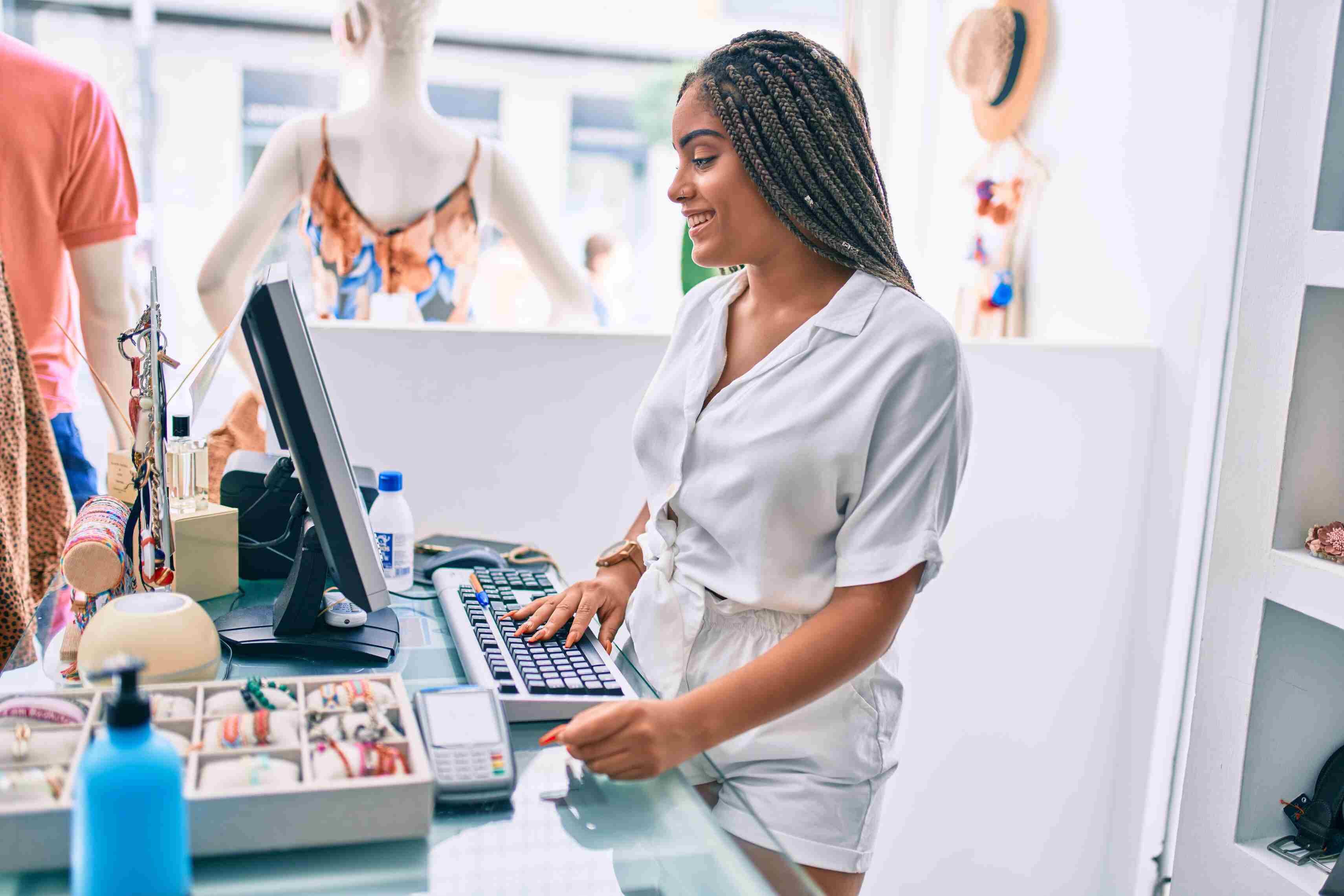 Smiling woman with braided hair working at a retail store counter, using a computer keyboard with jewelry and a payment terminal on the glass counter beside her.