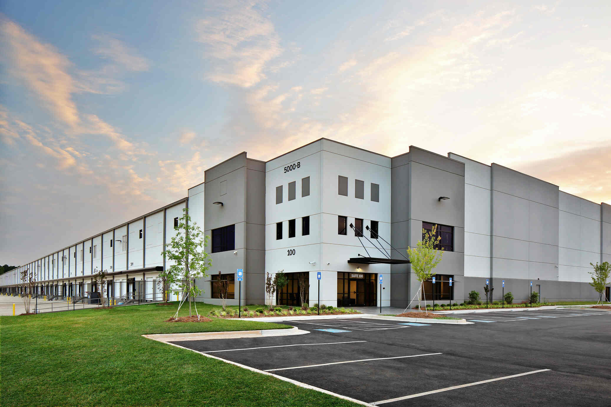 Large modern commercial warehouse building with gray and white exterior, multiple loading docks along the side, small trees planted outside, and an empty parking lot in front under a partly cloudy sky at sunset.