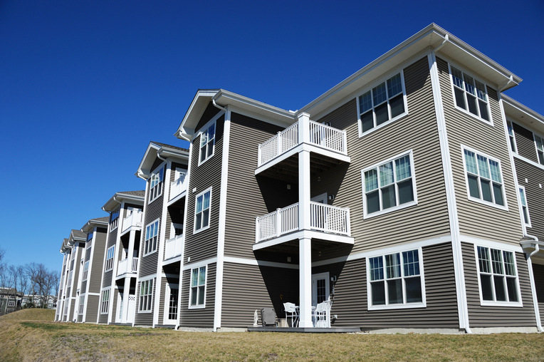 Exterior view of a modern multi-family apartment building with beige siding, white trim, and multiple balconies under a clear blue sky.