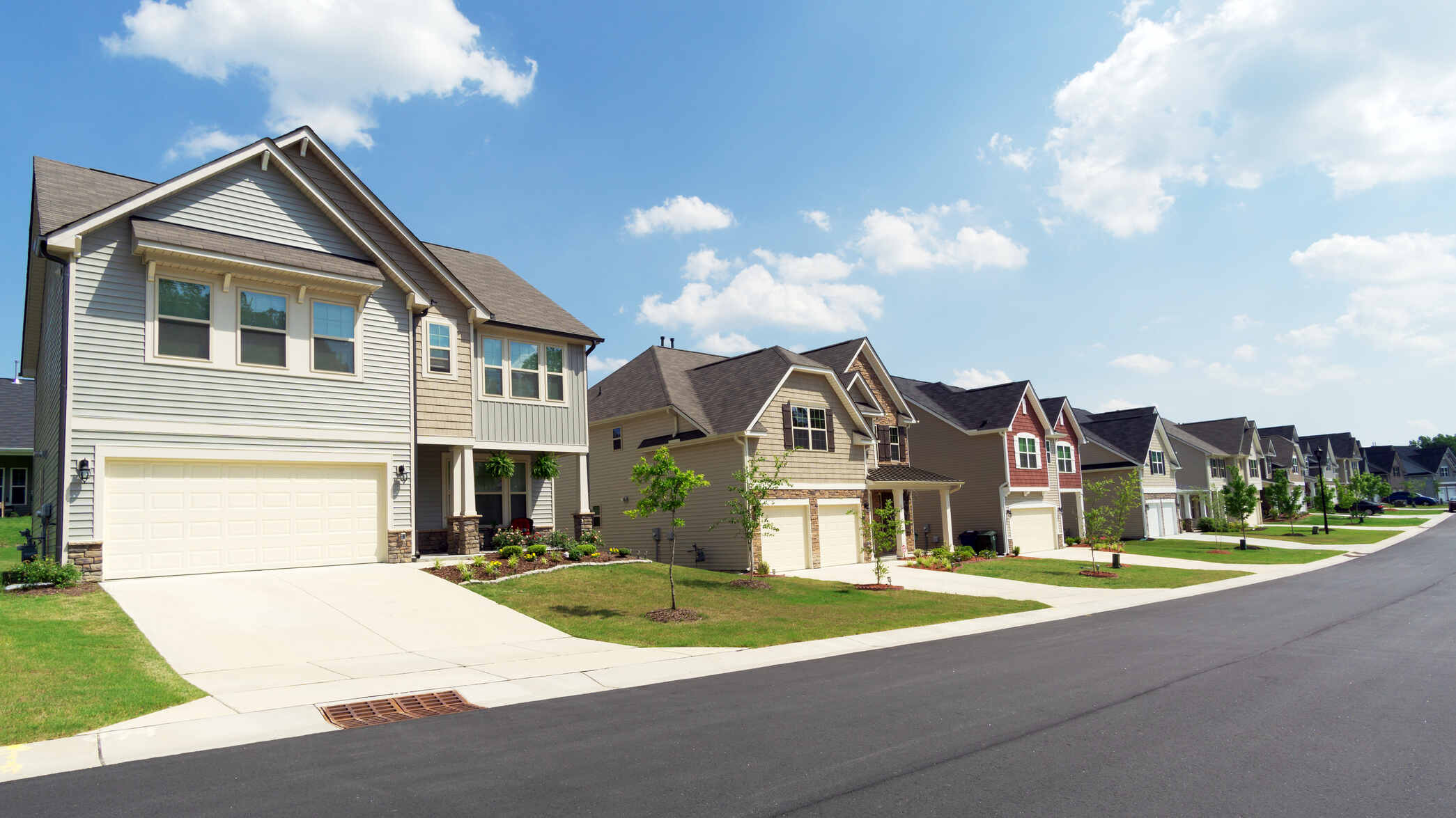 Row of modern suburban two-story houses with garages, small front lawns, and young trees along a clean asphalt street under a blue sky with scattered clouds.