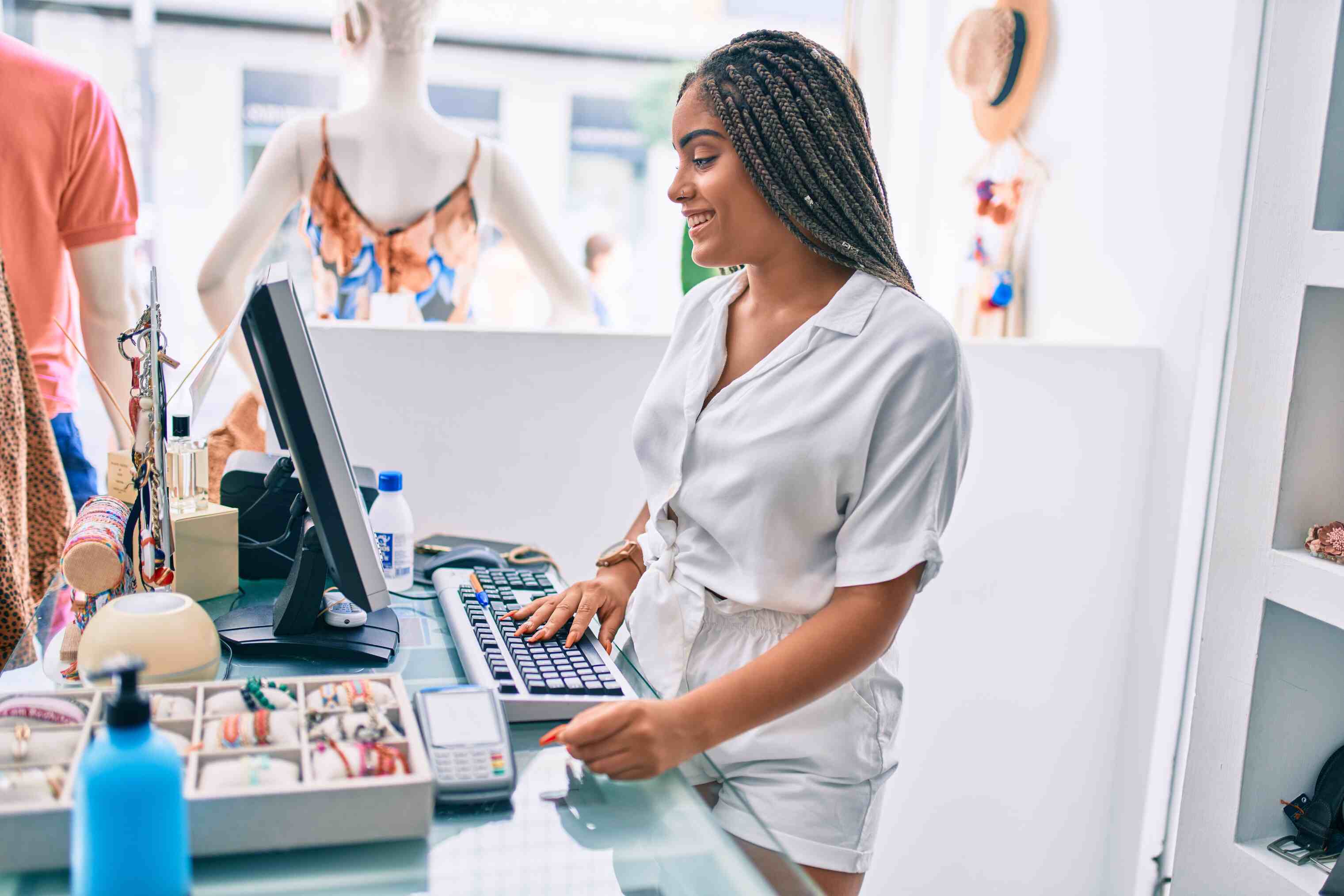 Smiling woman with braided hair wearing a white outfit standing at a glass counter in a retail store, typing on a keyboard with various jewelry displayed nearby.