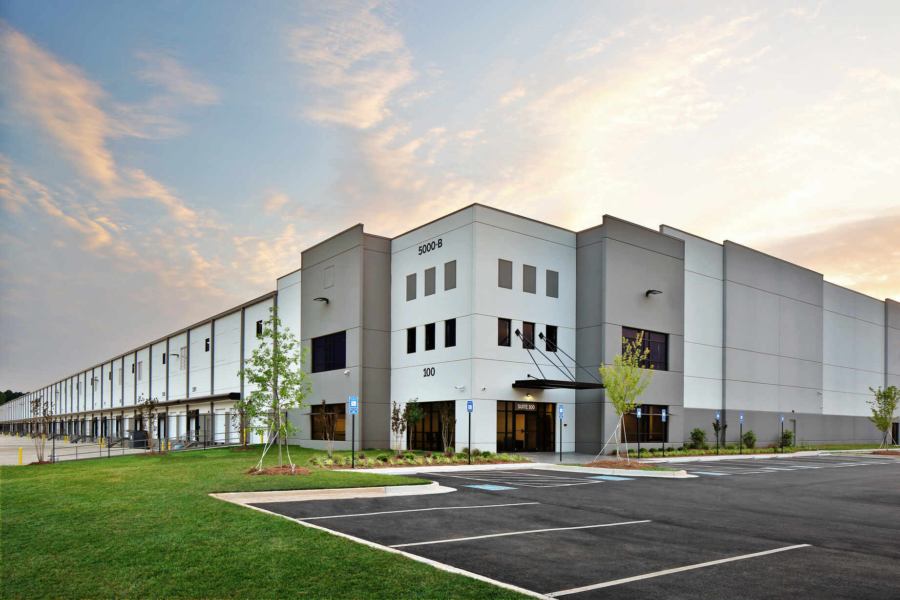 Modern commercial warehouse building with gray and white exterior and large windows under a partly cloudy sky during sunset, surrounded by parking spaces and landscaped grass with young trees.