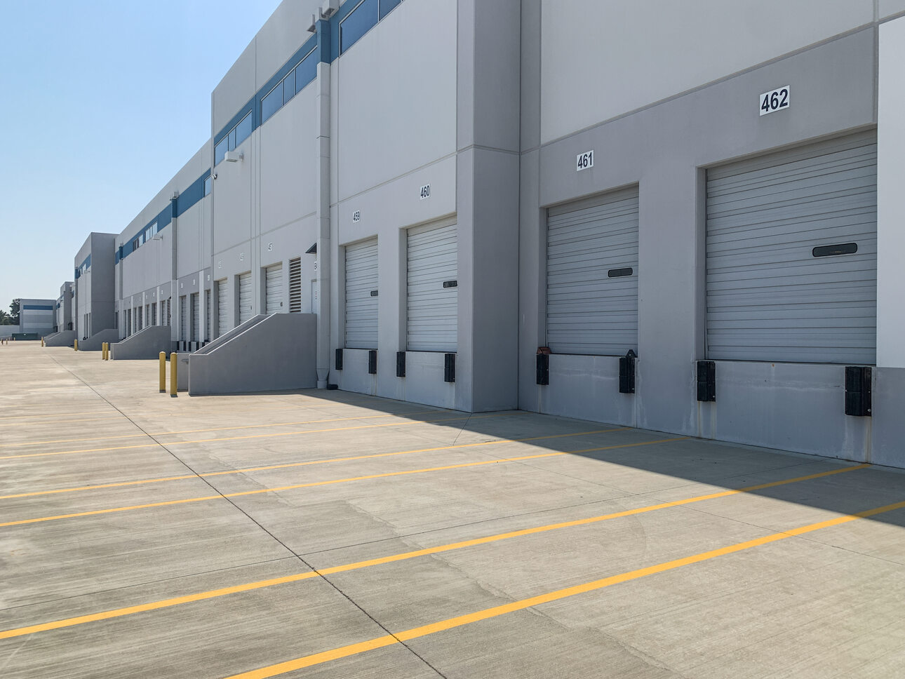 Exterior view of an industrial warehouse building with multiple numbered loading dock doors and yellow parking lines on the concrete ground under a clear blue sky.