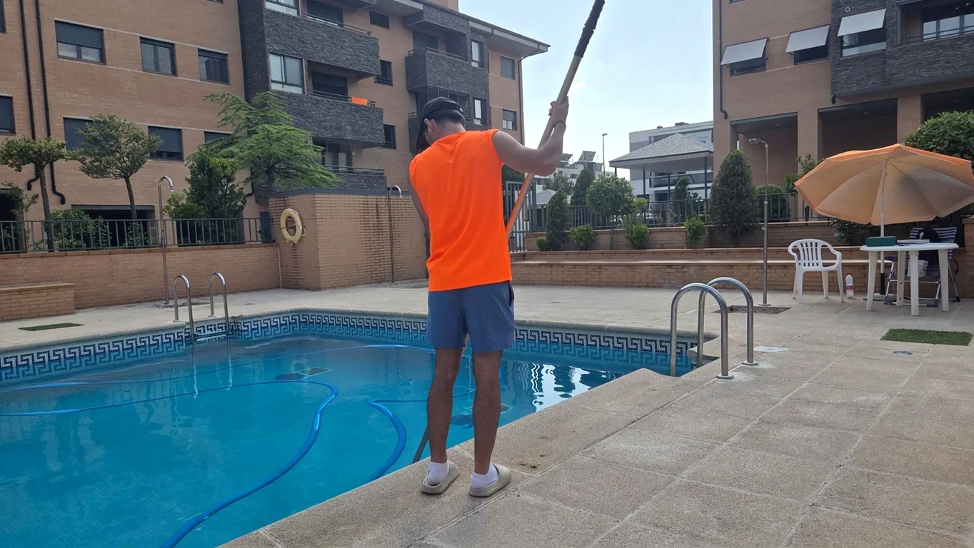 Person in an orange sleeveless shirt, blue shorts, white shoes, and black cap cleaning an outdoor swimming pool with a long-handled pool brush, surrounded by brick buildings, patio tiles, a white plastic chair, and a table with an orange umbrella.