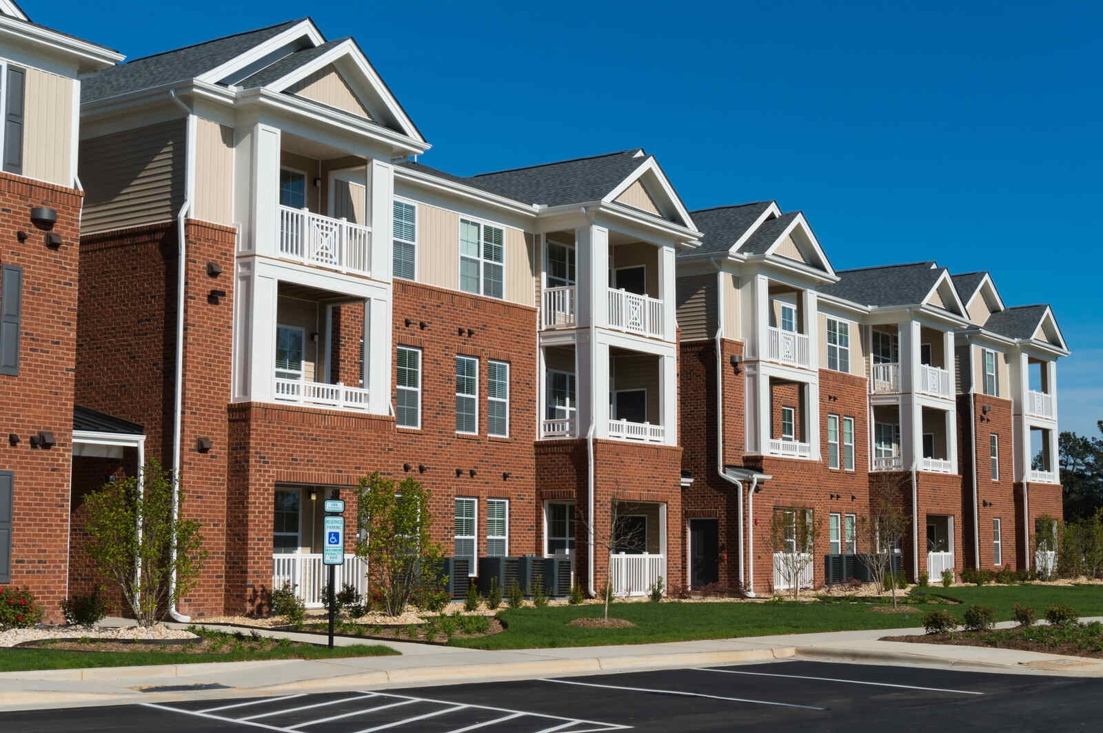 Modern three-story apartment building made of red brick and beige siding with multiple balconies, white railings, and a clear blue sky above.