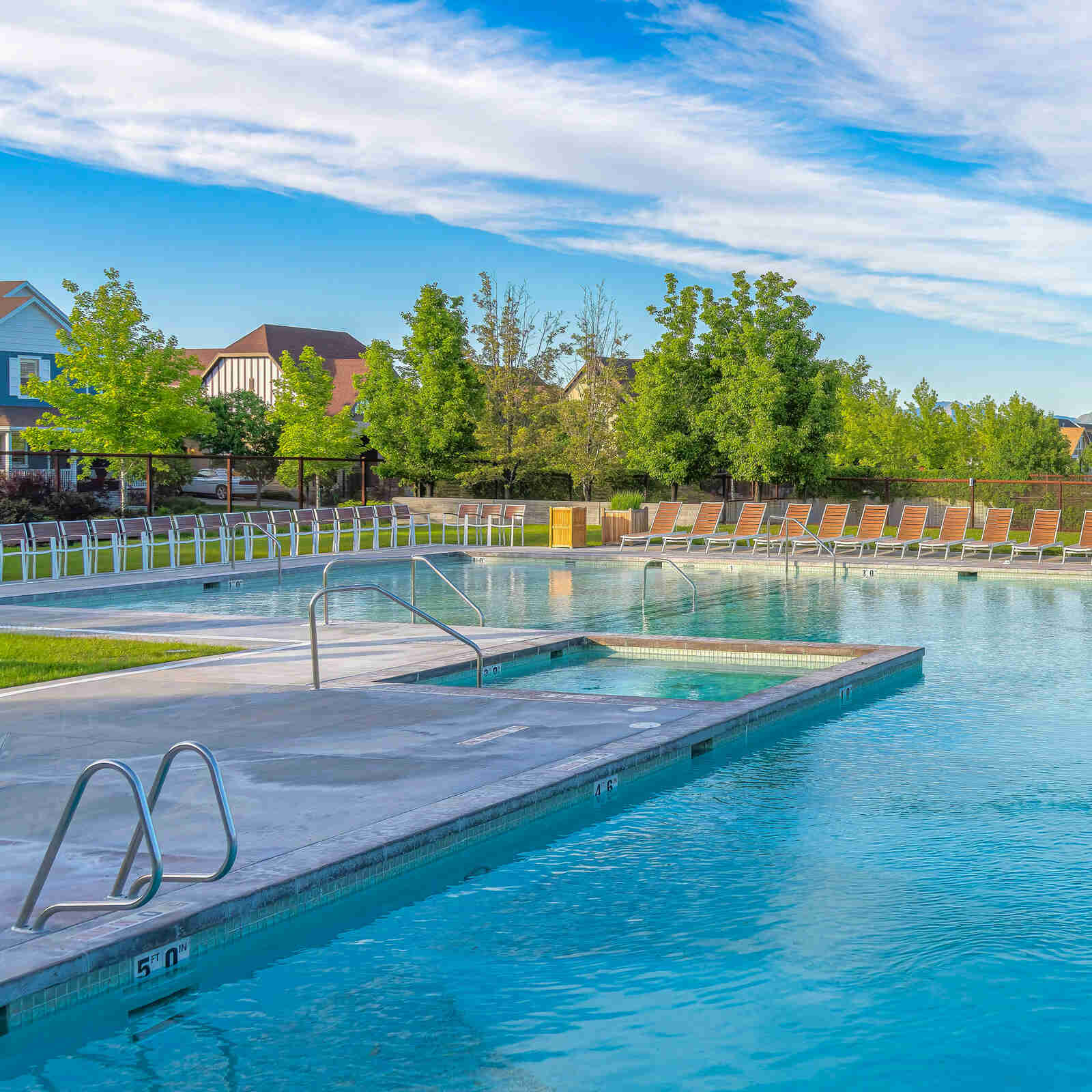 Outdoor swimming pool with clear blue water, an integrated hot tub, metal handrails, and a row of wood and metal lounge chairs on a concrete deck, surrounded by green trees and residential houses under a partly cloudy blue sky.
