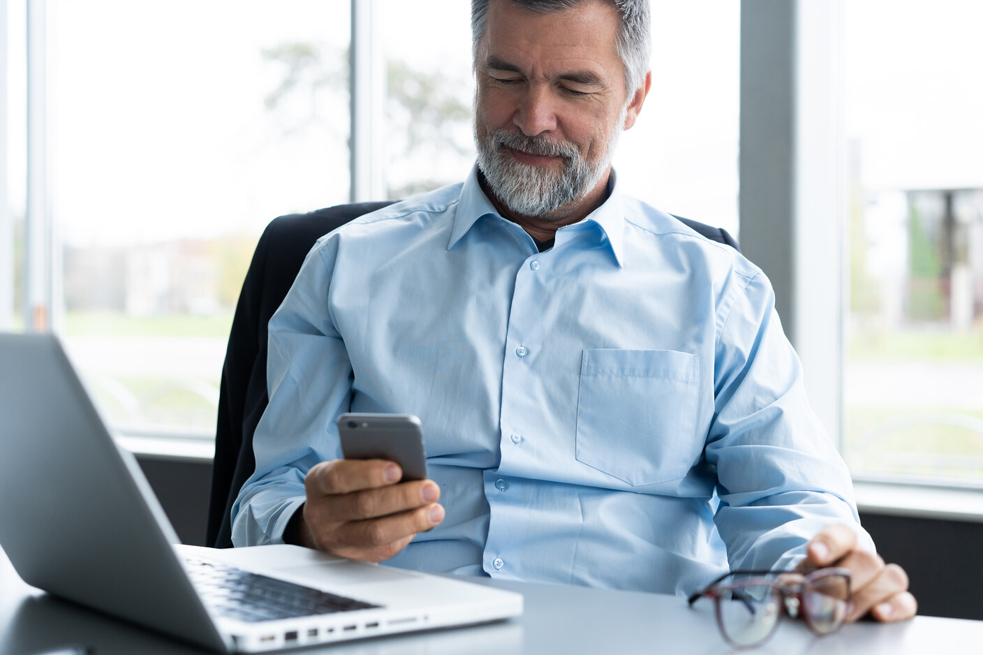 Middle-aged man with gray hair and beard sitting at a desk, looking at his smartphone with a slight smile, with a laptop open in front of him and holding eyeglasses in his other hand.