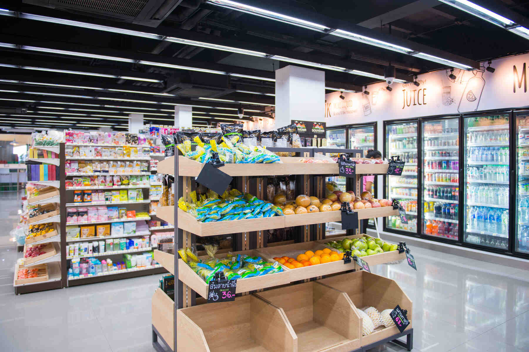 Interior of a modern grocery store with a wooden display rack featuring packaged snacks, oranges, green apples, and pomelos, with refrigerated sections for drinks visible in the background.