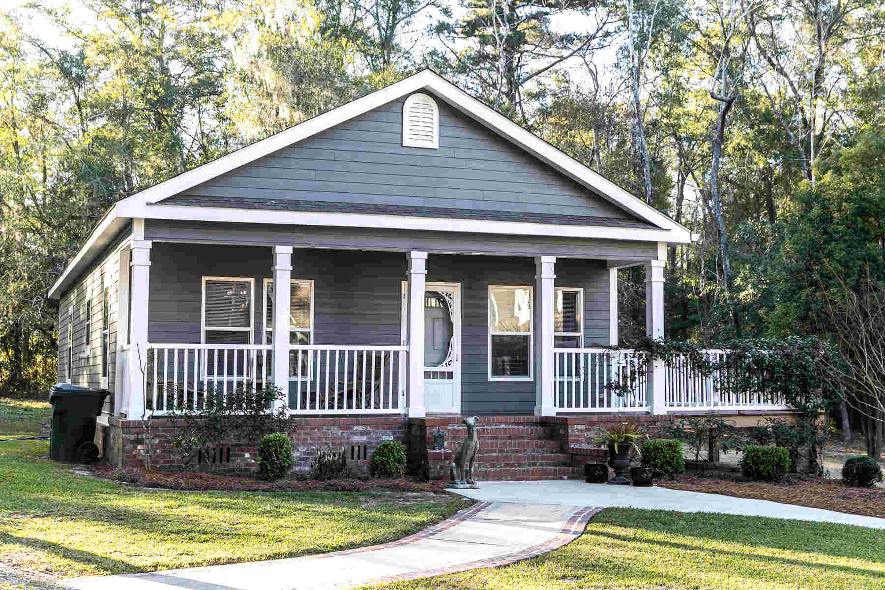 Single-story gray house with white trim and a covered porch supported by four white columns, surrounded by shrubs and trees with a curved concrete walkway leading to the porch steps.