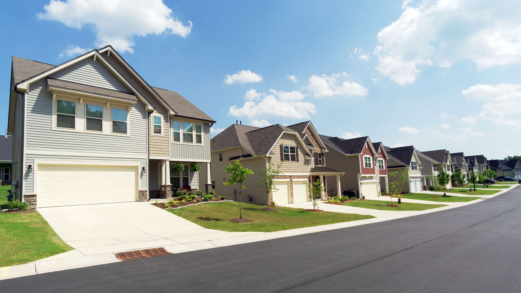 Residential street with a row of modern suburban two-story houses featuring garages, neatly trimmed lawns, young trees, and a clear sky with scattered clouds.