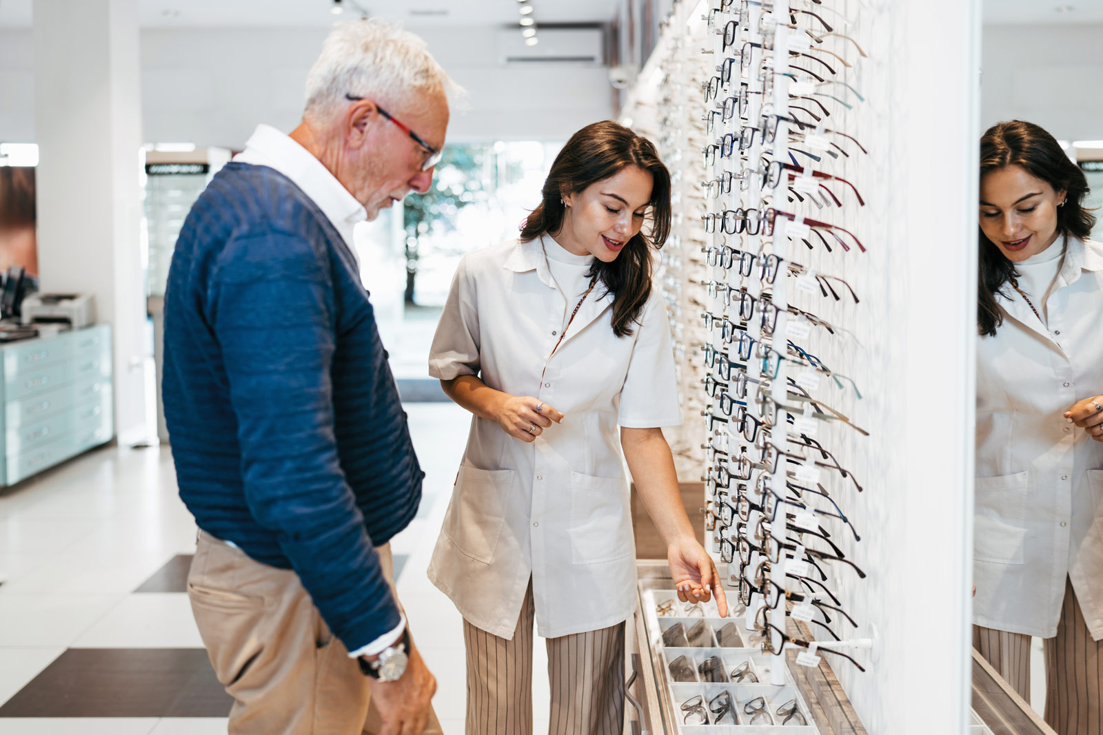 An older man wearing glasses and a blue sweater stands next to a woman in a white coat at an optical store, where she points to eyeglasses displayed in a case under a wall filled with various eyeglass frames.