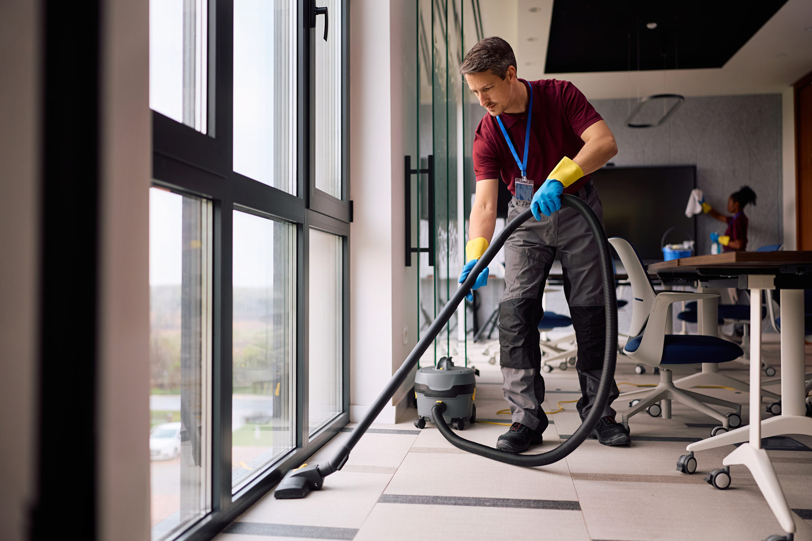 A janitor vacuuming the floor along a large glass window in a modern office while a colleague cleans a wall-mounted screen in the background.