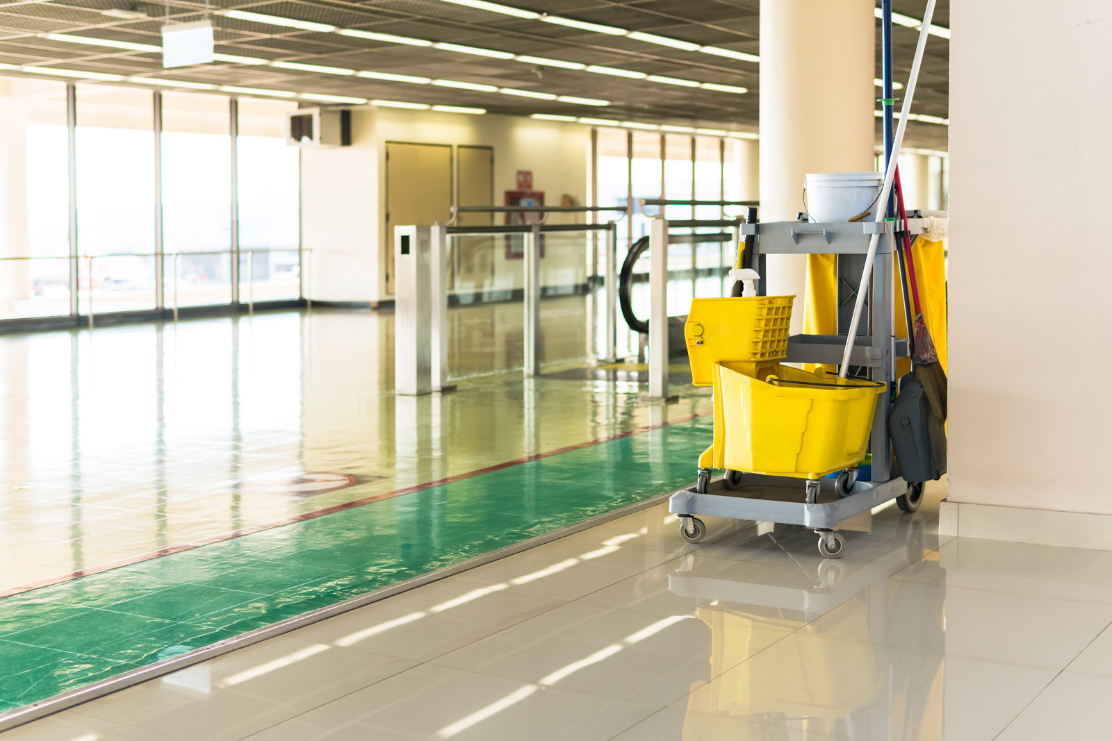 Janitorial cleaning cart with yellow mop buckets, mop, broom, and cleaning supplies in a brightly lit, polished indoor hallway near glass windows and doors.