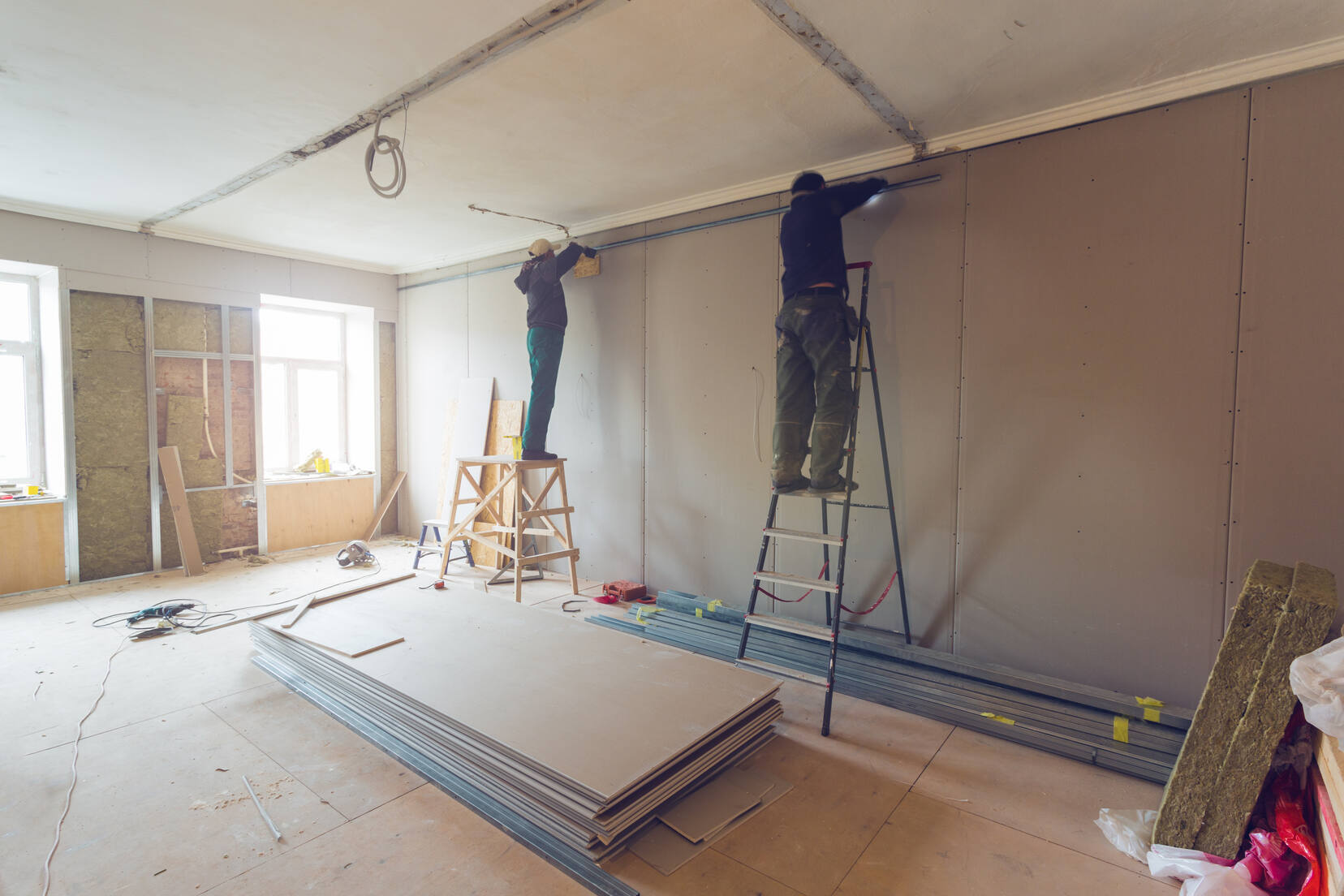 Two workers standing on ladders installing drywall in a room under renovation, with construction materials and insulation scattered around.