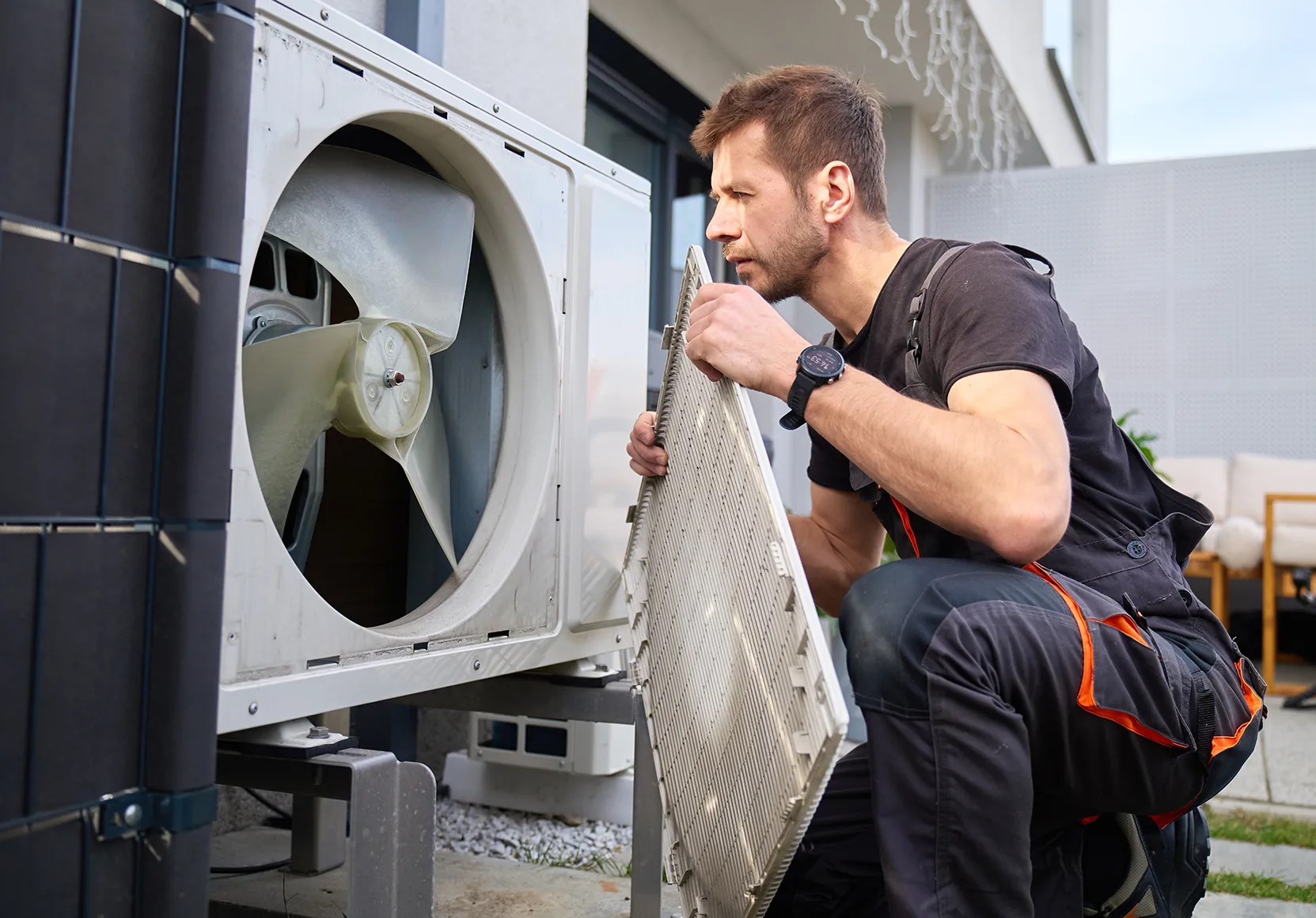 A technician in black and orange workwear inspects an outdoor air conditioning unit's fan while holding a removed grill panel, in a residential setting.
