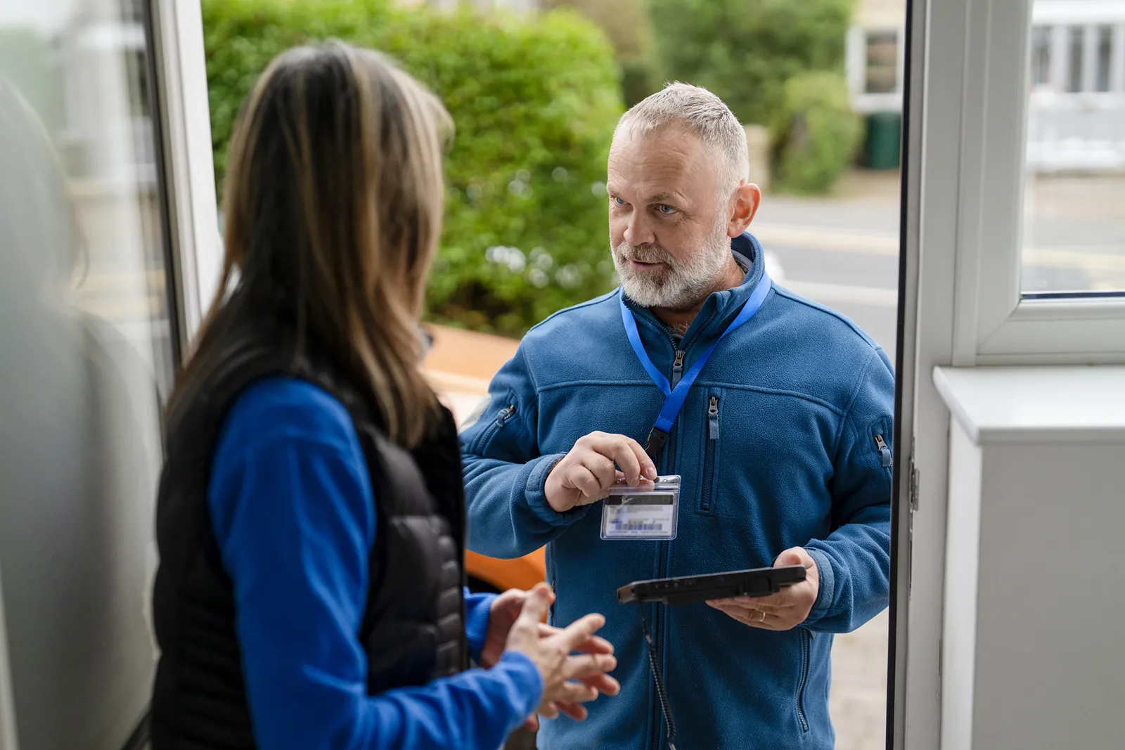 Middle-aged man in a blue fleece jacket showing an ID badge on a lanyard to a woman standing inside a doorway, holding a tablet and talking to her outdoors.