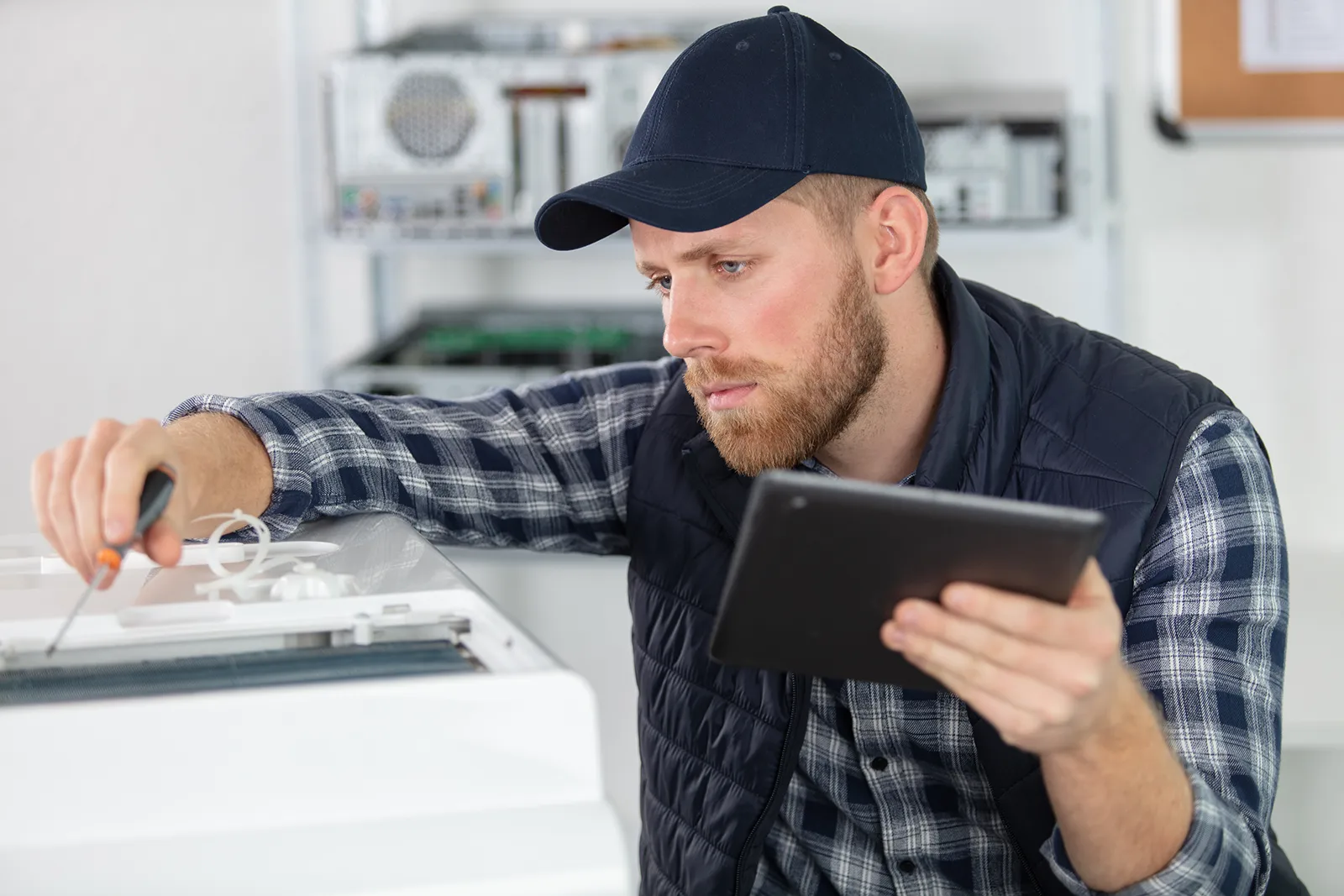 A man in a navy cap and plaid shirt with a dark vest uses a screwdriver to repair a white appliance while holding a black tablet.