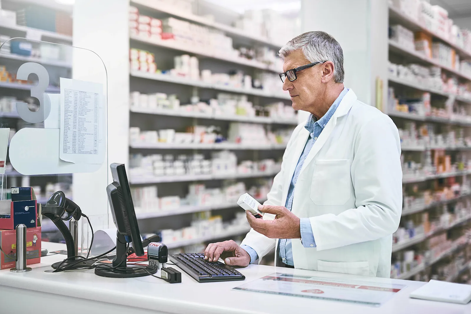 A pharmacist wearing glasses and a white lab coat stands behind a counter in a pharmacy, holding a medication box and typing on a computer keyboard with shelves of medicines in the background.