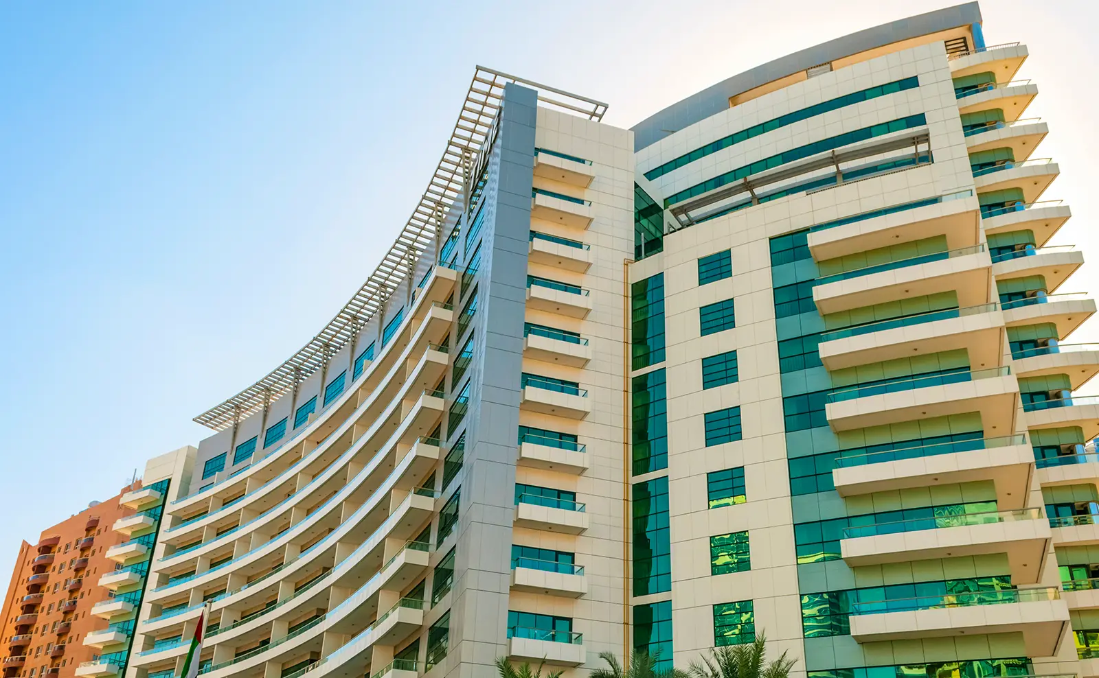 Modern multi-story building with curved facade, green-tinted glass windows, and balconies under a clear blue sky.