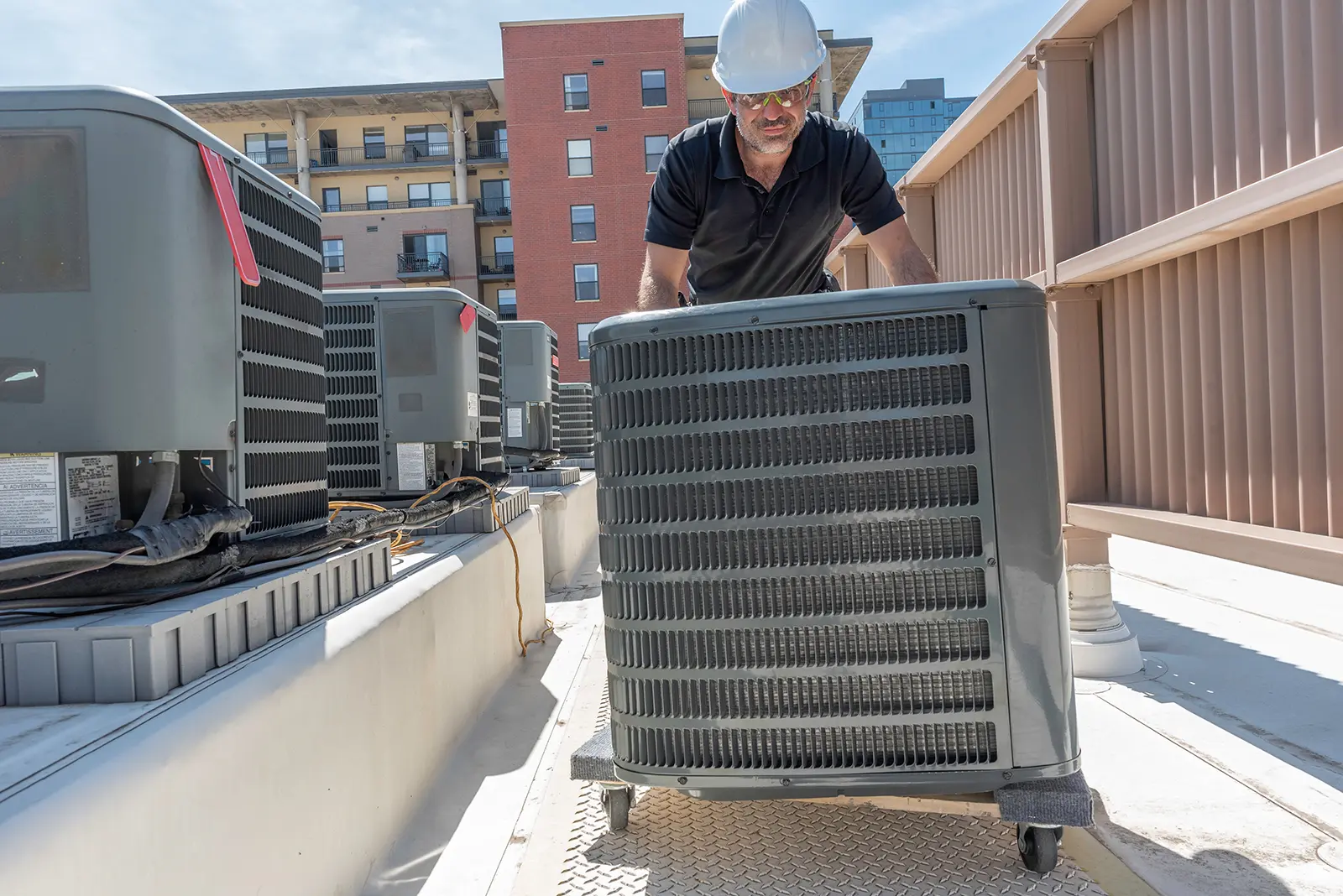 Technician wearing a white hard hat, safety glasses, and black shirt moving a large HVAC unit on a wheeled dolly on a rooftop with apartment buildings in the background.