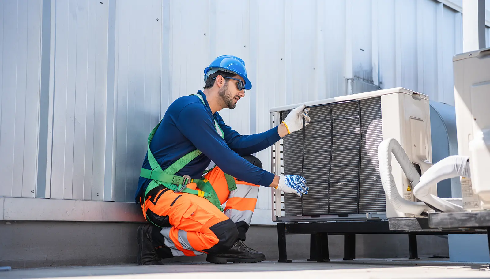 Technician wearing a blue hard hat, safety glasses, gloves, and orange reflective pants kneeling while inspecting an outdoor air conditioning unit beside a metal building.