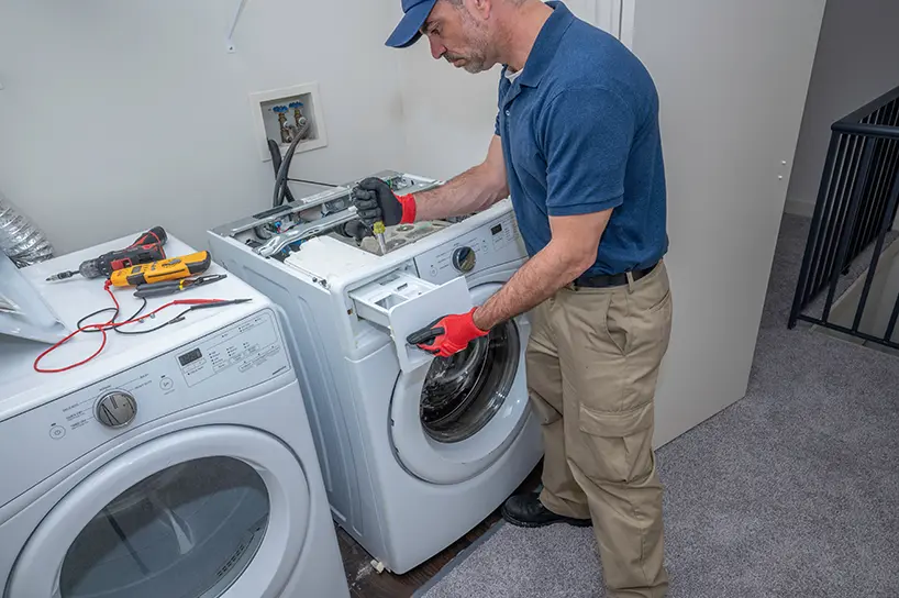 Technician in a blue shirt and cap repairing a front-loading washing machine, using a screwdriver while standing next to a dryer with tools on top in a laundry room.