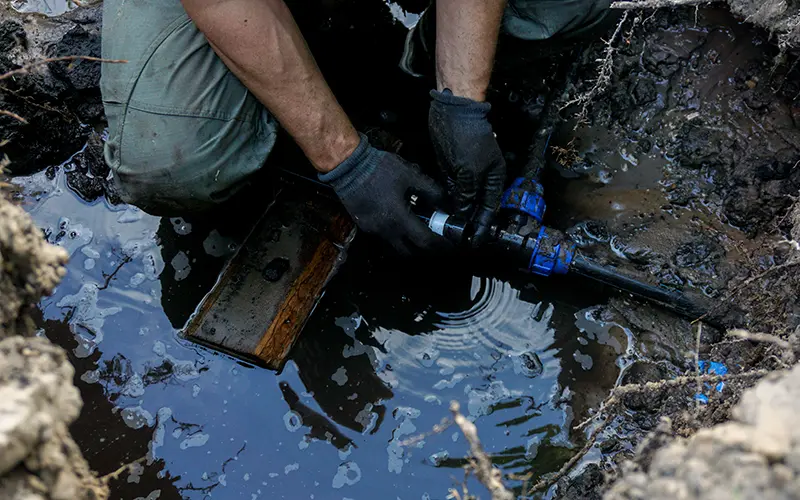 Person wearing gloves and green pants repairing a black and blue water pipe in muddy water and soil outdoors.