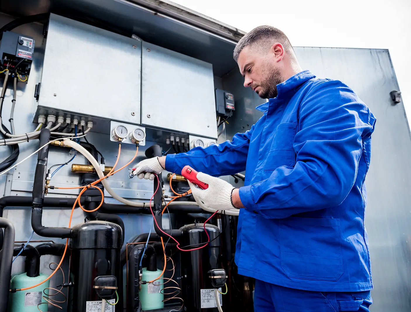 Technician in blue uniform and white gloves testing or repairing HVAC equipment with pressure gauges and connected pipes.
