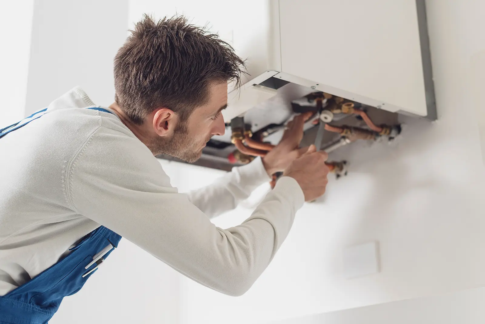 A man in a white long-sleeve shirt and blue overalls repairs pipes underneath a wall-mounted boiler using a wrench.