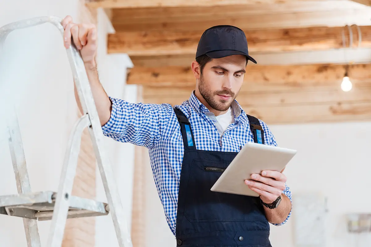 Man in a black cap and overalls holding a tablet in one hand and standing next to a metal ladder inside a room with wooden beams on the ceiling and a hanging light bulb.