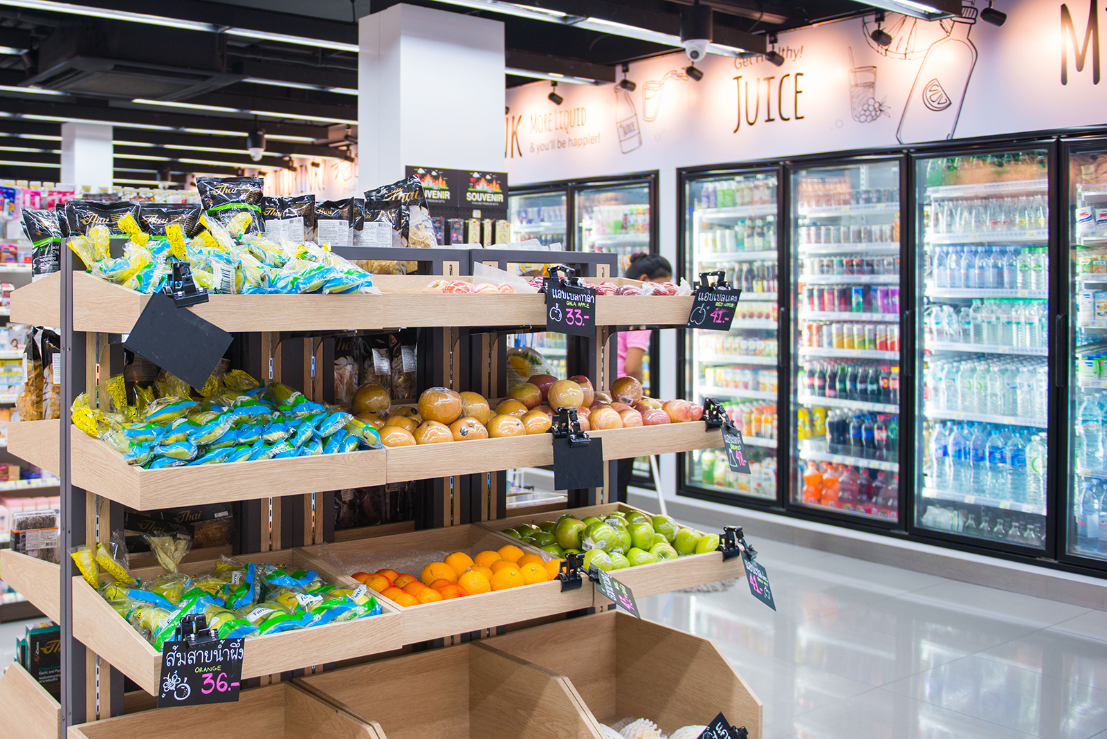 Interior of a supermarket produce section with wooden shelves displaying packaged snacks, oranges, apples, and other fruits, with refrigerated beverage coolers in the background.