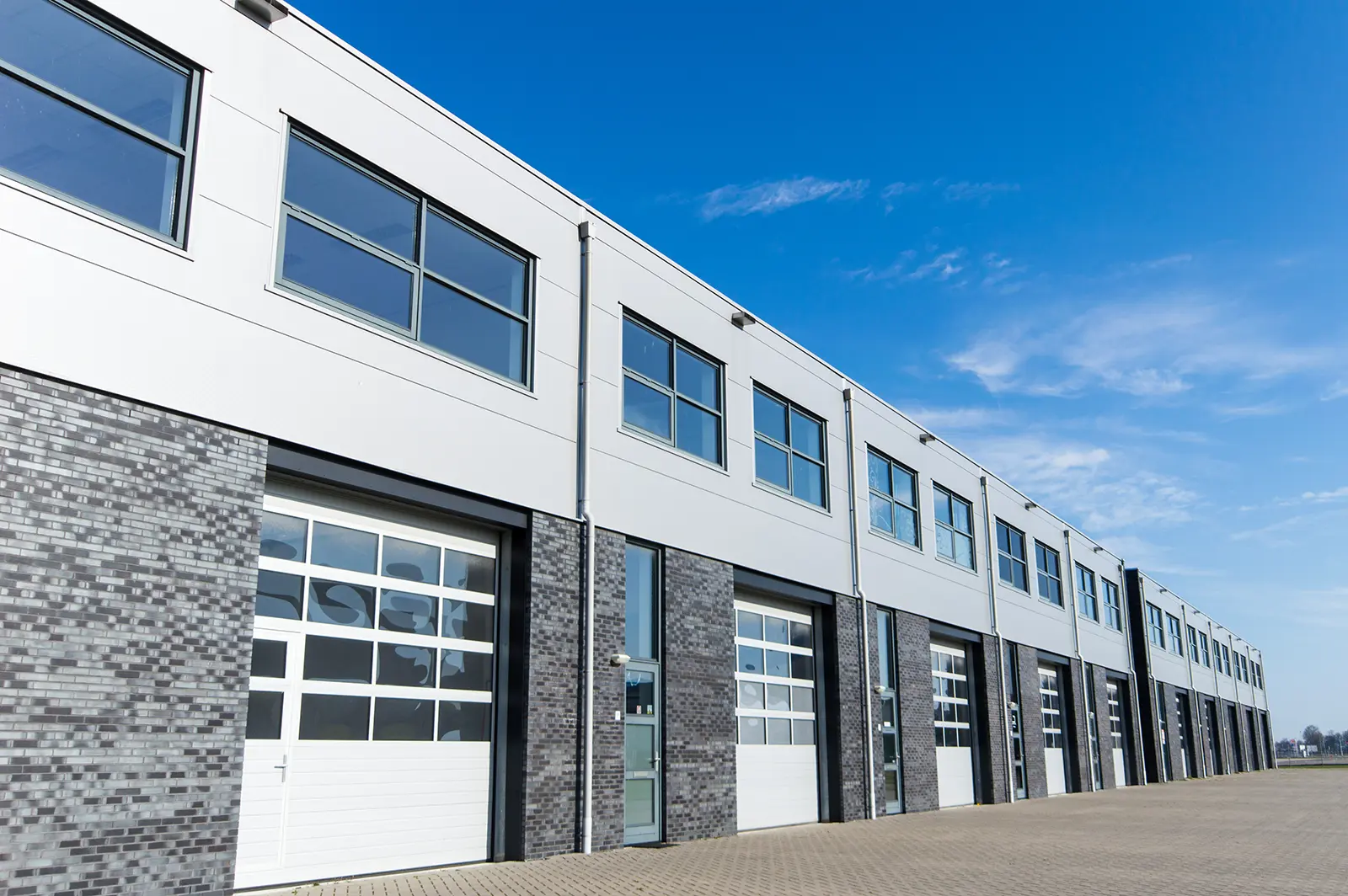 Row of modern industrial warehouse units with large white garage doors and upper-level windows under a clear blue sky.