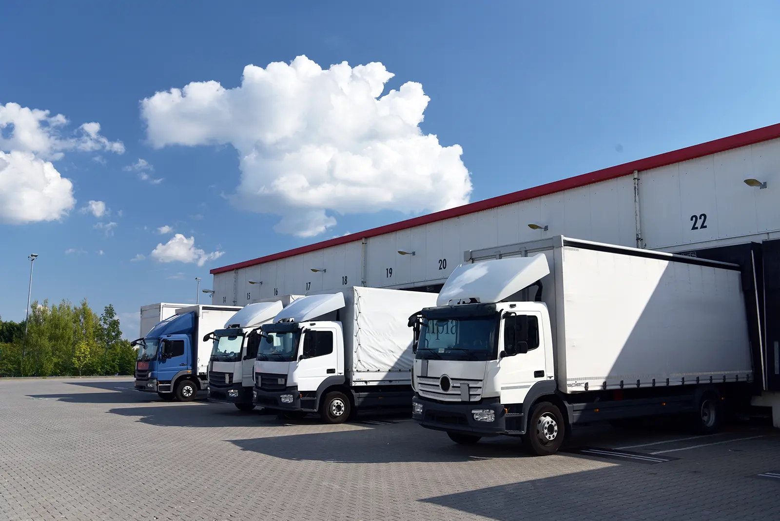 Four trucks, three white and one blue, parked in front of a warehouse loading dock under a partly cloudy sky.