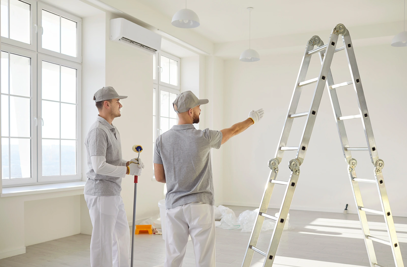 Two painters in gray shirts and white pants inside a bright room with large windows, one holding a paint roller and the other pointing towards a metal ladder, suggesting preparation for painting.