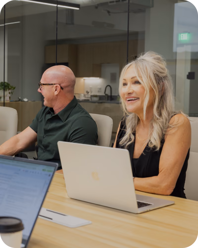 Two people sit at a conference table with laptops, smiling and engaged in a modern office meeting room with glass walls and a kitchen area in the background.