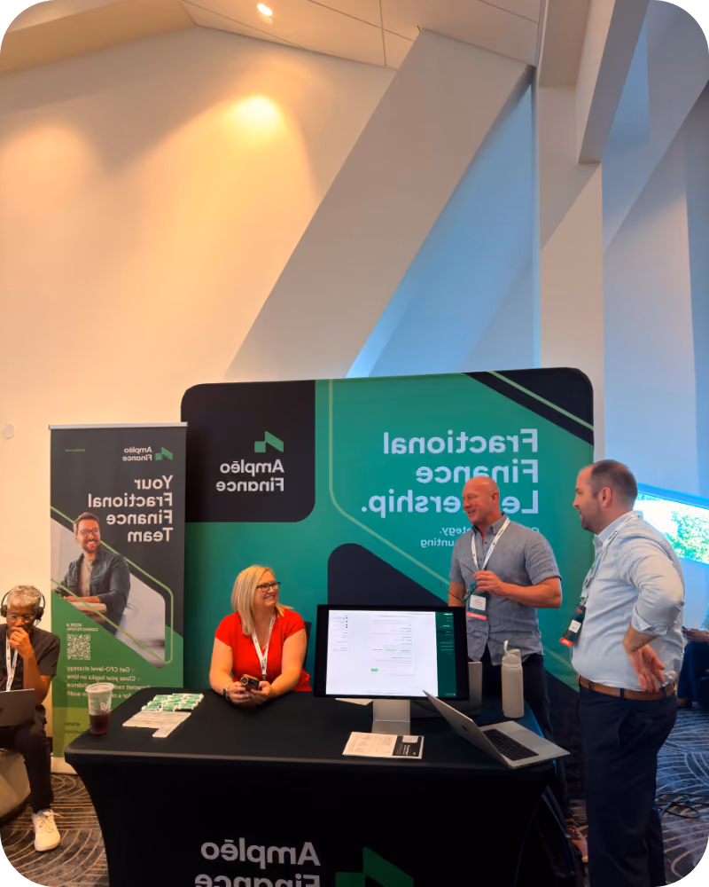 Three people at a trade show booth for Ampleo Finance; one woman in a red shirt sits behind the table with a computer monitor and promotional materials while two men stand talking nearby; a banner and backdrop display the company name and phrases about fractional finance leadership.