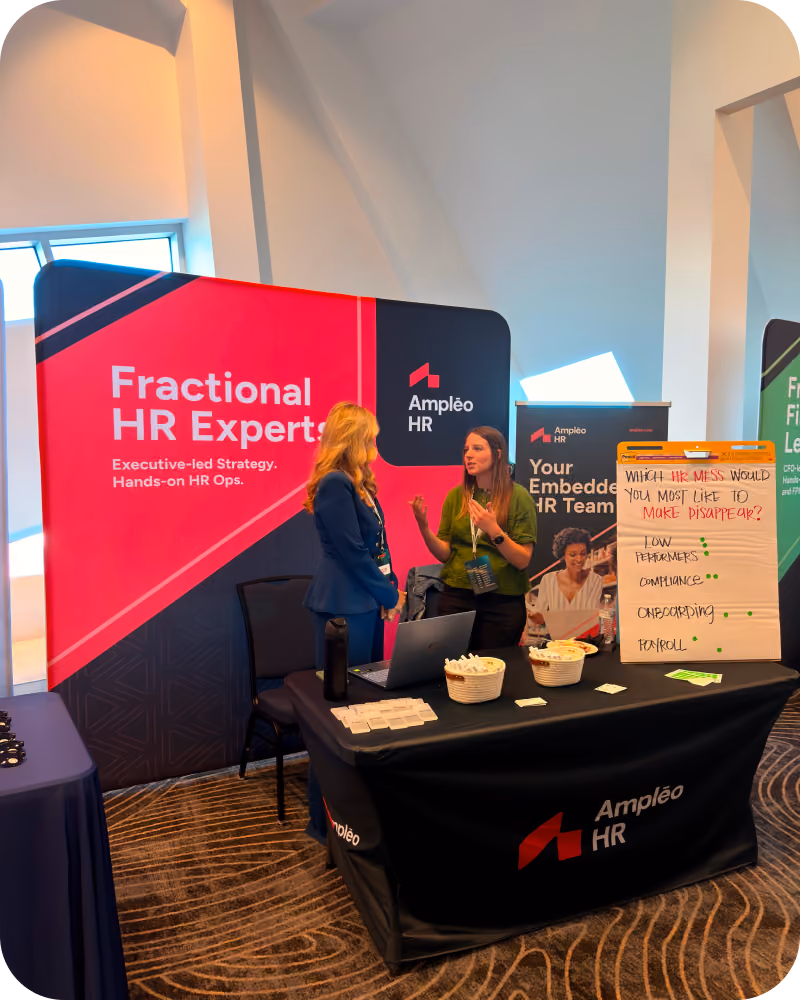 Two women engaged in conversation at an Ampleo HR booth with a large pink and black banner reading 'Fractional HR Experts' and a table covered with promotional materials, snacks, and a laptop. A flip chart nearby asks, 'Which HR mess would you most like to make disappear?' with options for low performers, compliance, onboarding, and payroll marked with green dots.