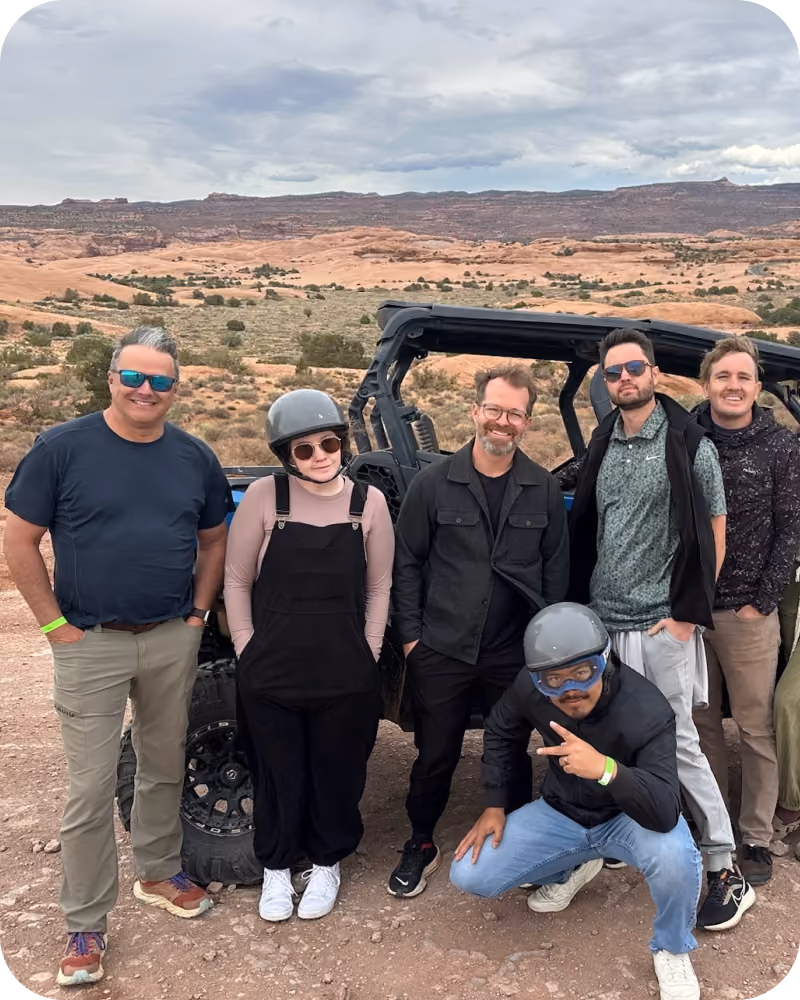 Six people posing outdoors in a desert landscape with red rock formations in the background, standing and crouching beside a black off-road vehicle, some wearing helmets and sunglasses.