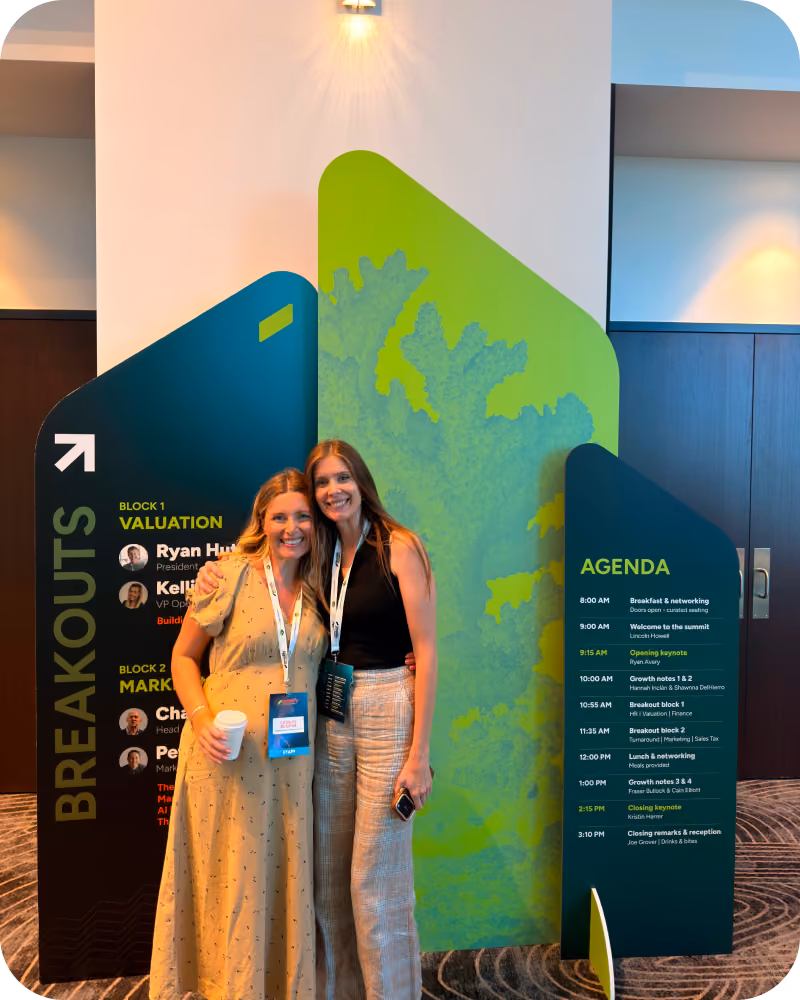 Two smiling women wearing conference badges stand arm in arm in front of large green and black signs displaying a conference breakout and agenda schedule. The signs list session blocks on valuation and marketing and the day's agenda from 8:00 AM to 3:10 PM.